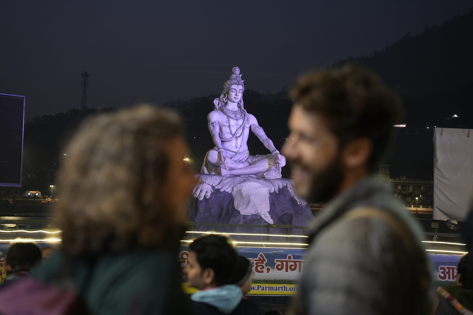 The floodlit Shiva statue at Parmarth Niketan at night, with two visitors out of focus in the foreground