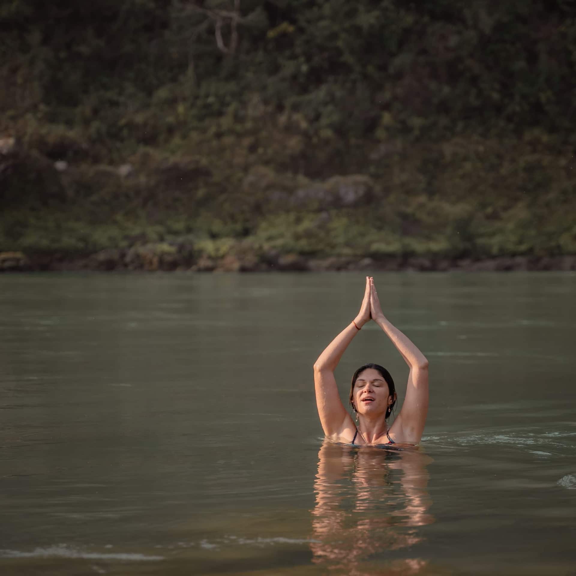 A woman standing chest-deep in the Ganga with her eyes closed and her palms pressed together above her head