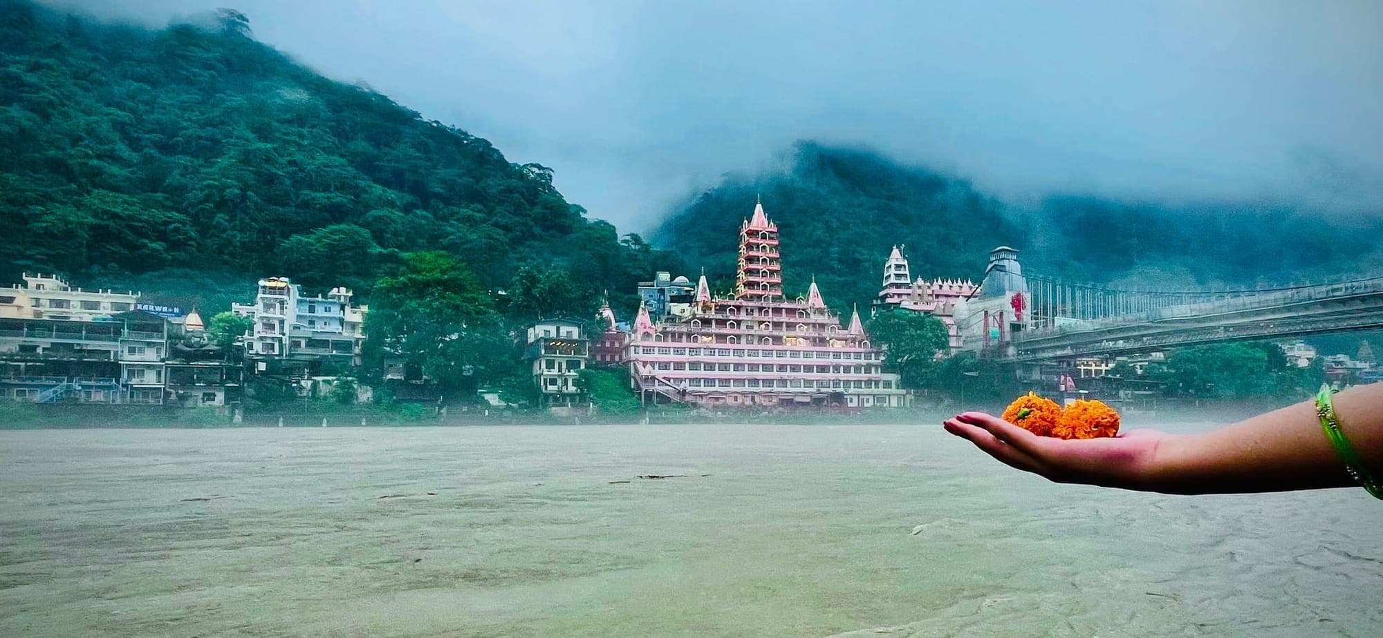Marigold flower offering held out over the Ganga, with Trayambakeshwar Temple, Laxman Jhula bridge and misty hills across the river