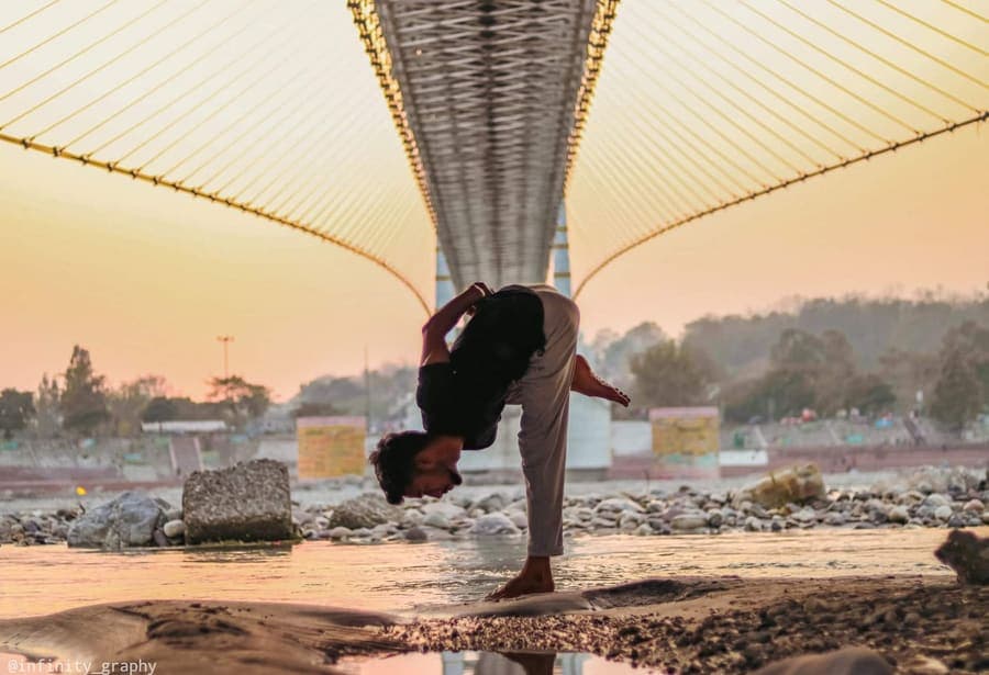 A man practising a standing forward-bend yoga pose on the banks of the Ganga beneath a cable bridge in Rishikesh at sunset