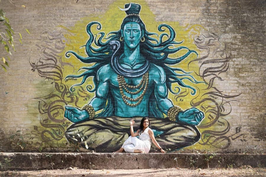 A woman in white sits beneath a large painted mural of a meditating Shiva on a brick wall at the Beatles Ashram in Rishikesh