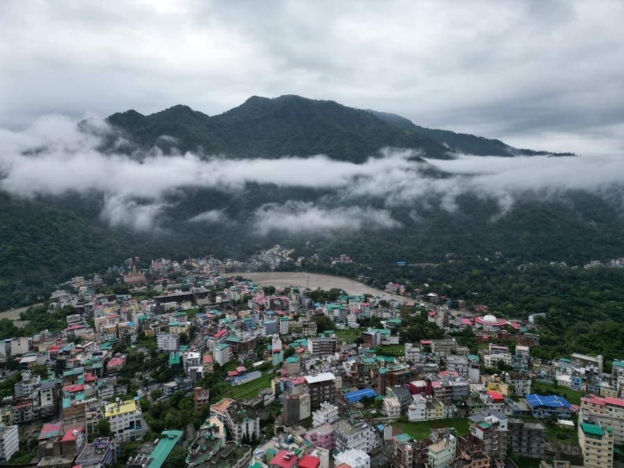 Aerial view of Rishikesh town rooftops beside a bend in the Ganga river, with forested hills and low monsoon cloud behind