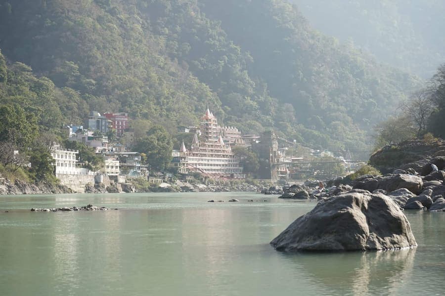Hazy morning view along the pale green Ganga in Rishikesh toward a tiered pink temple and hillside buildings