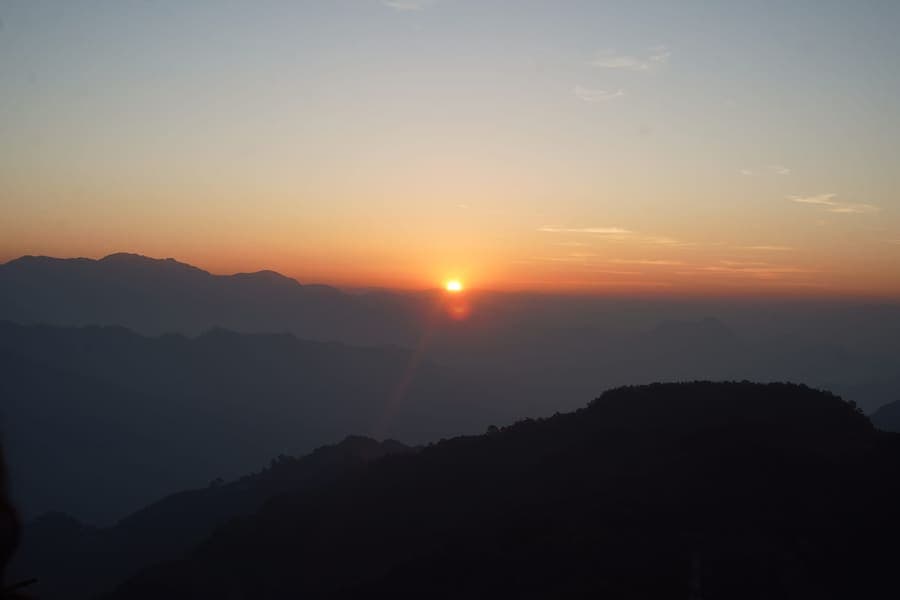 The sun low on the horizon above layered Himalayan foothill ridges, with an orange sky and dark forested slopes in silhouette