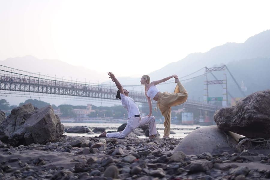 Two people practising yoga poses on the stony bank of the Ganga in Rishikesh, with a suspension bridge and misty hills behind