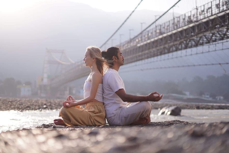 A woman and a man meditating back to back beside the Ganga river in Rishikesh, with suspension bridges behind them