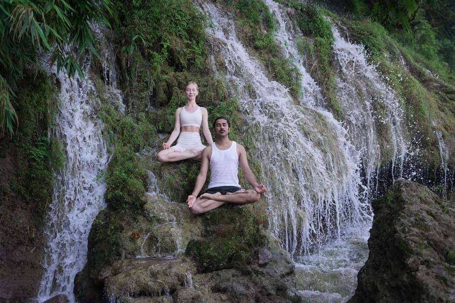 Two people meditating in lotus posture on mossy rocks in front of a waterfall near Rishikesh