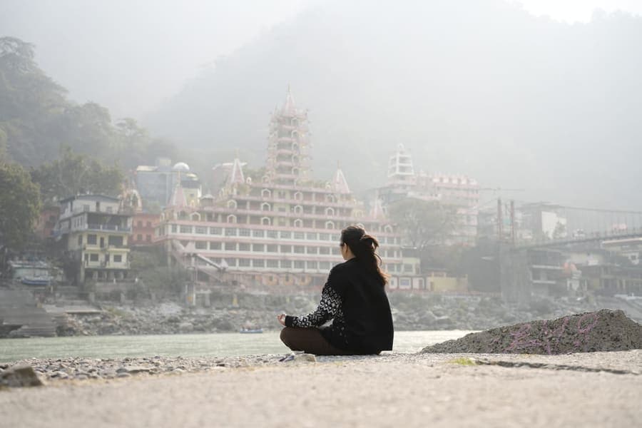 Woman meditating on the Ganga riverbank facing the tiered Trimbakeshwar temple and misty hills at Laxman Jhula, Rishikesh