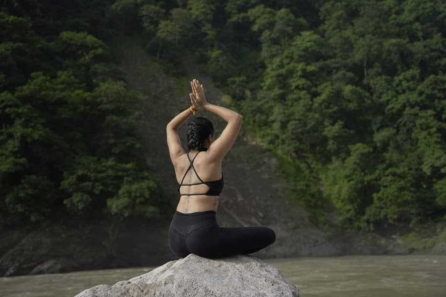 Woman seated cross-legged on a boulder beside the Ganga with palms joined overhead, facing a forested hillside
