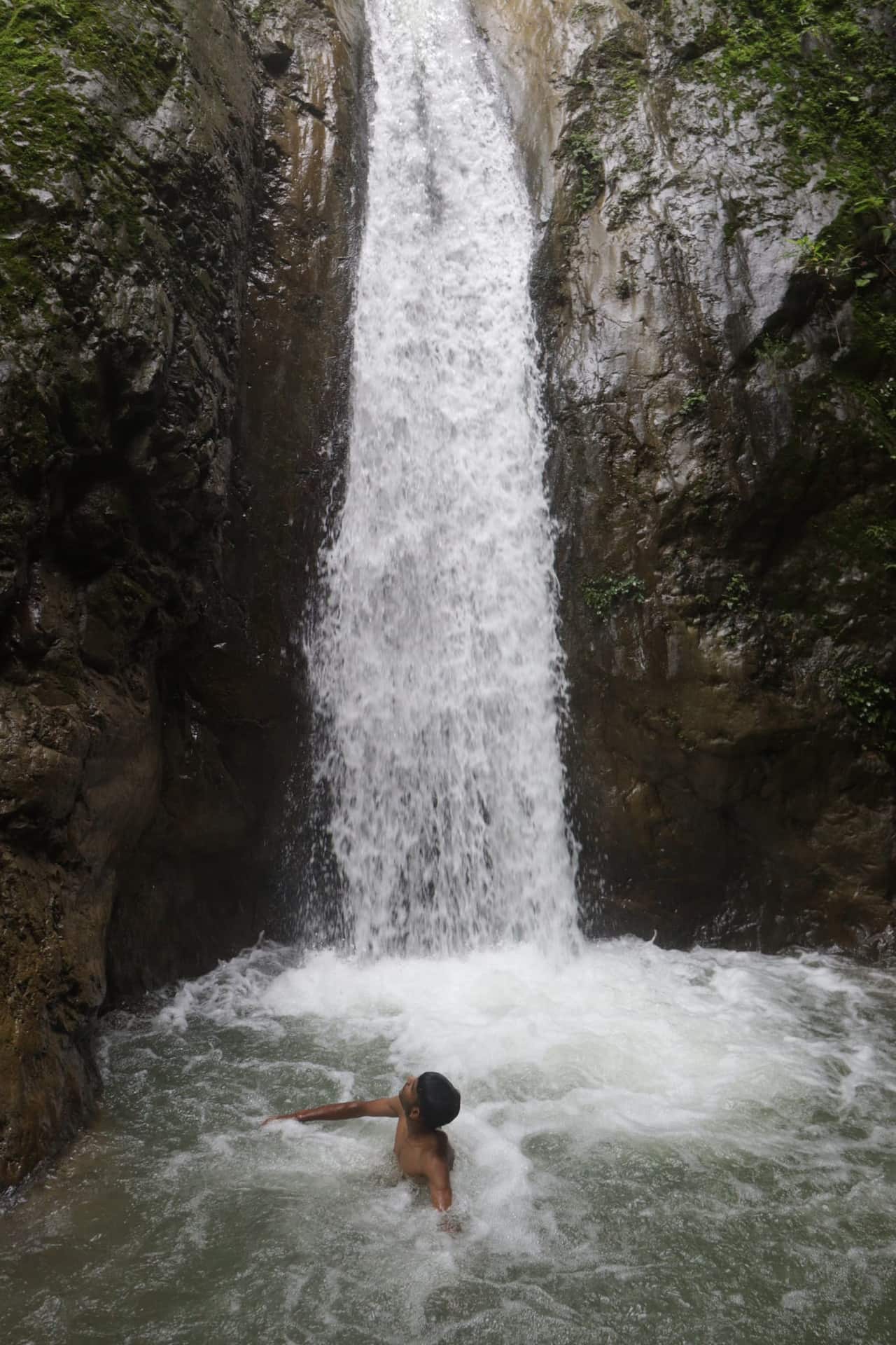 A person standing in the plunge pool of a tall waterfall in a rocky gorge near Rishikesh