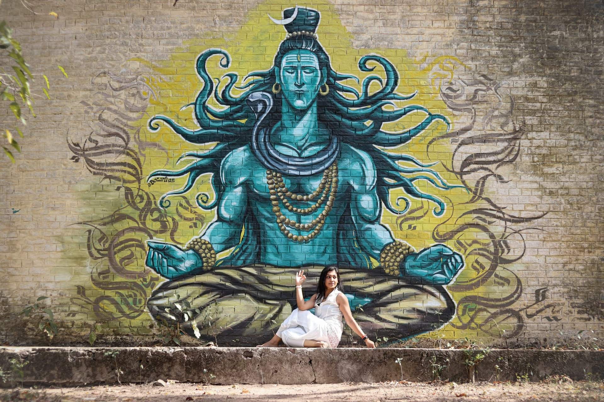 A woman in white sits beneath a large painted mural of a meditating Shiva on a brick wall at the Beatles Ashram in Rishikesh