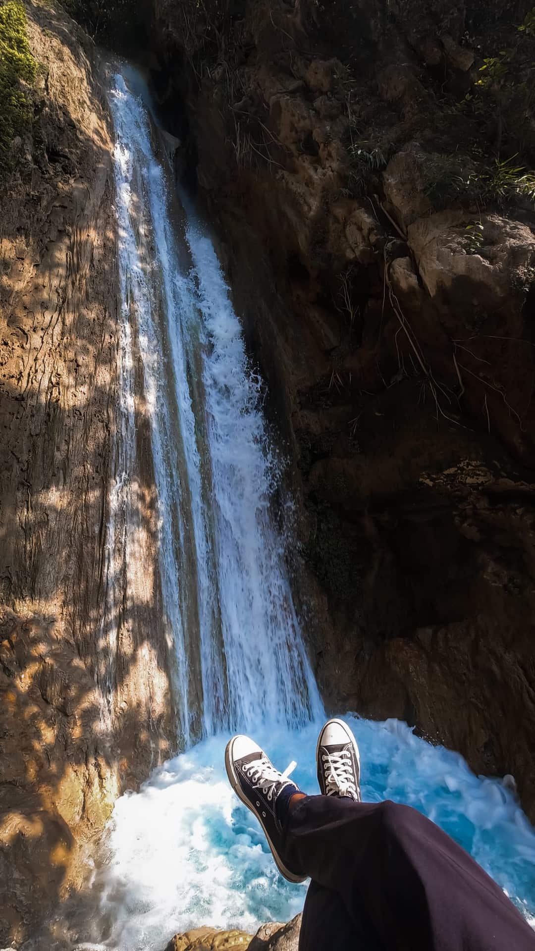 Legs in black sneakers dangling over the edge of a waterfall in Rishikesh, with white water falling down a rock face into a turquoise pool