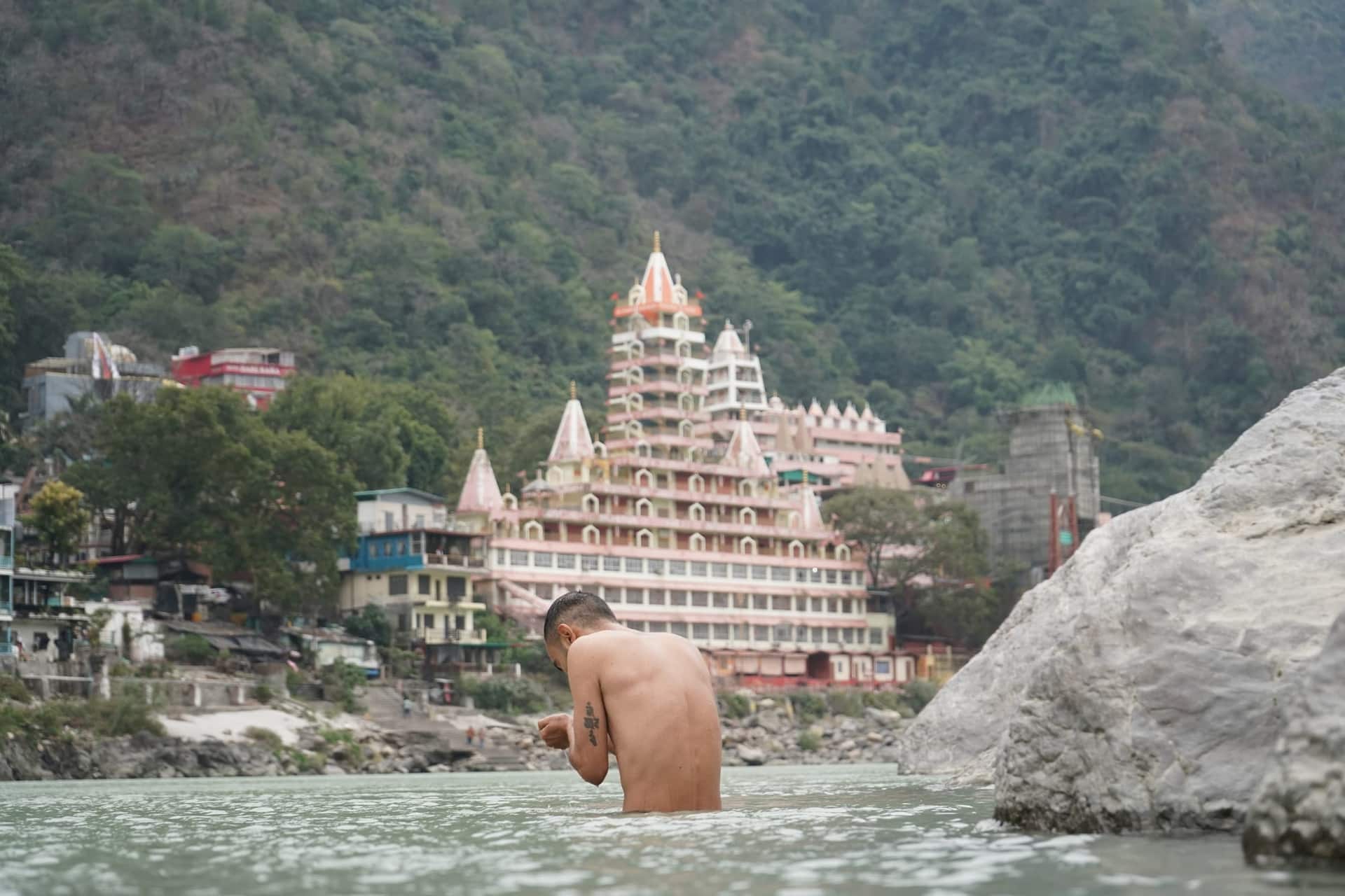 A man stands waist-deep in the Ganga with head bowed and hands joined, the tiered pink riverside temple of Rishikesh behind him.