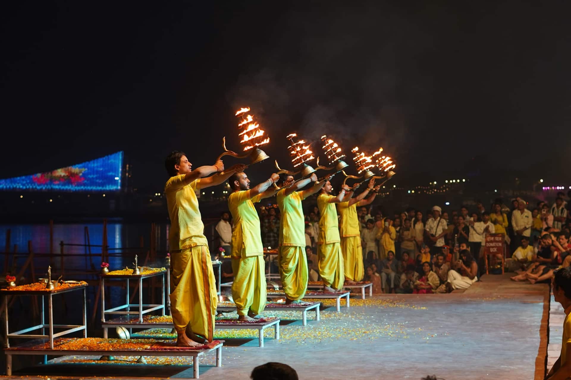 Priests in yellow robes raise tiered brass fire lamps in a line during an evening Ganga aarti ceremony at a riverside ghat, watched by a large crowd.