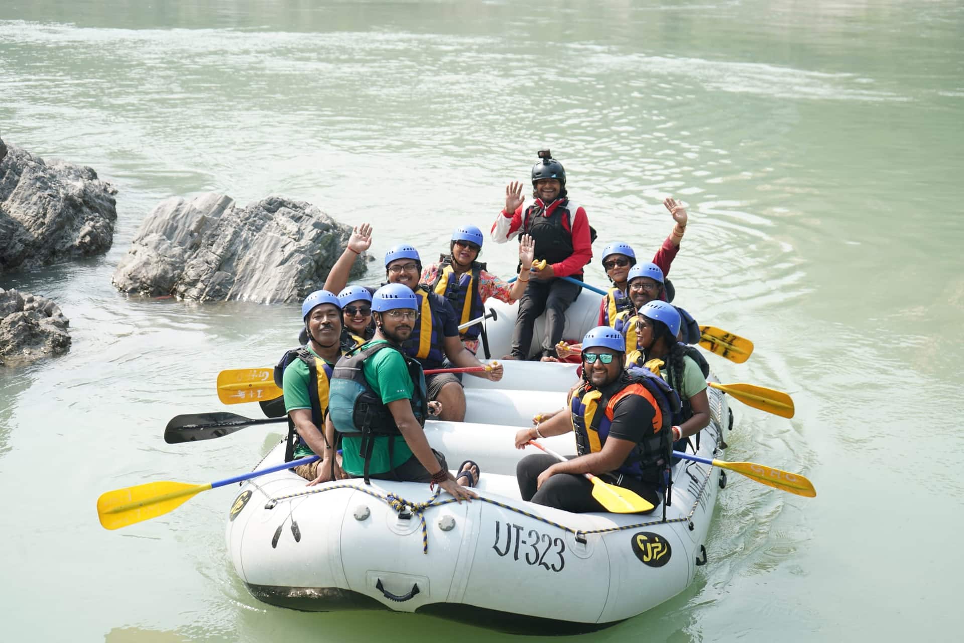 Group of rafters in blue helmets and life jackets wave from a white raft on the Ganga river near Rishikesh