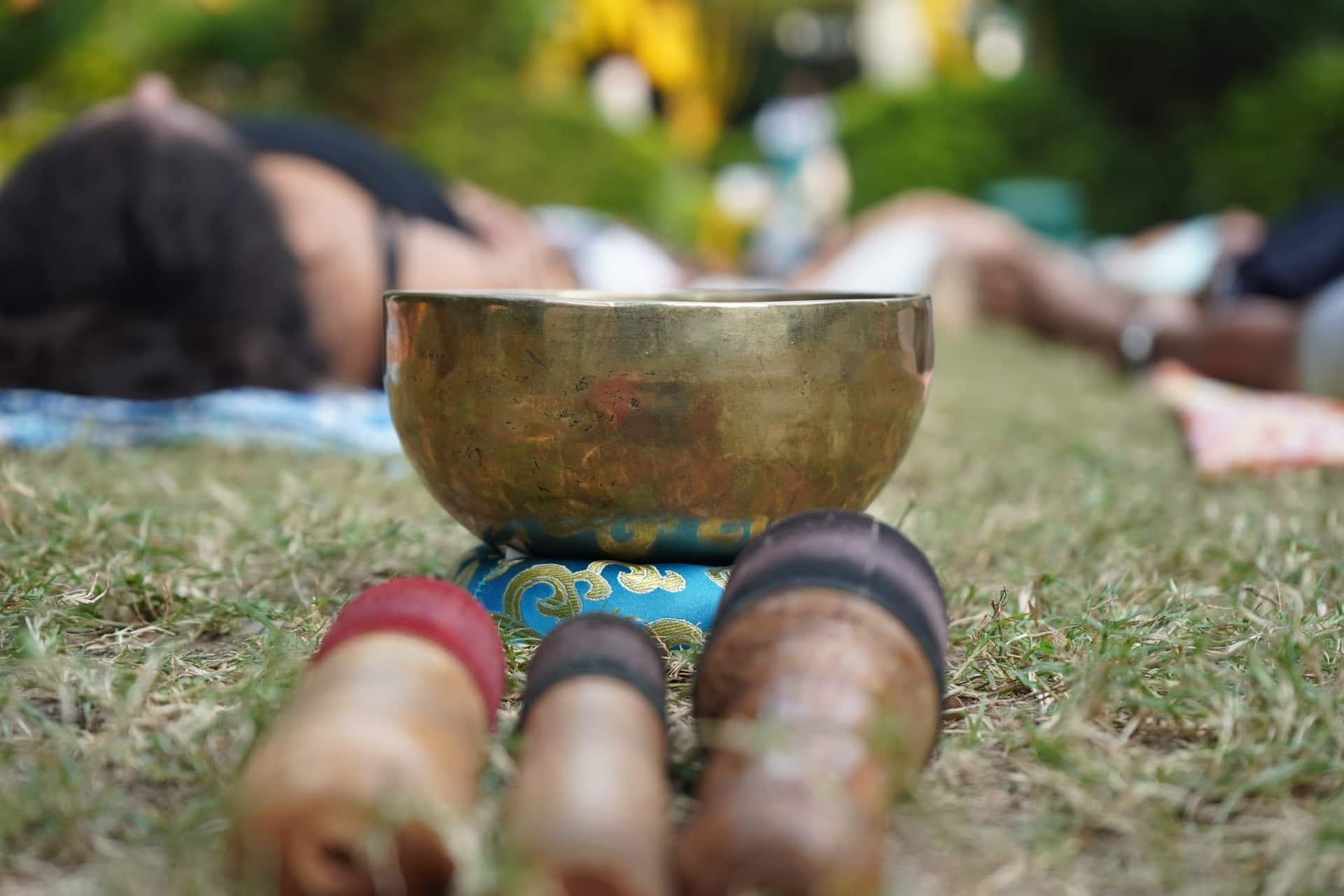 Brass singing bowl on a patterned cushion on grass, with people lying down for a sound healing session blurred in the background.