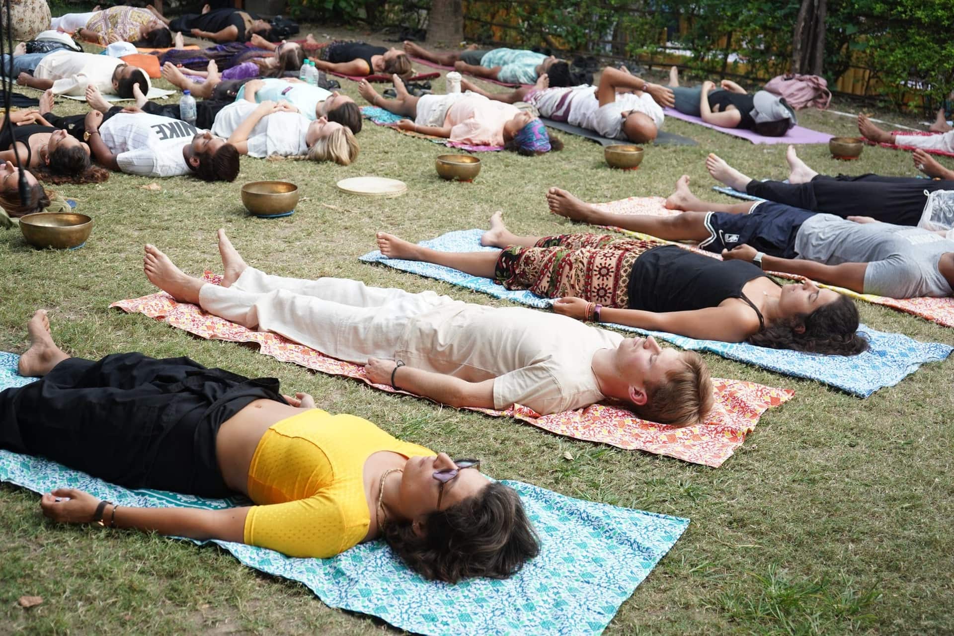 Around thirty people lying on printed cotton mats on a lawn during an outdoor sound healing and relaxation session, with brass singing bowls placed on the grass.