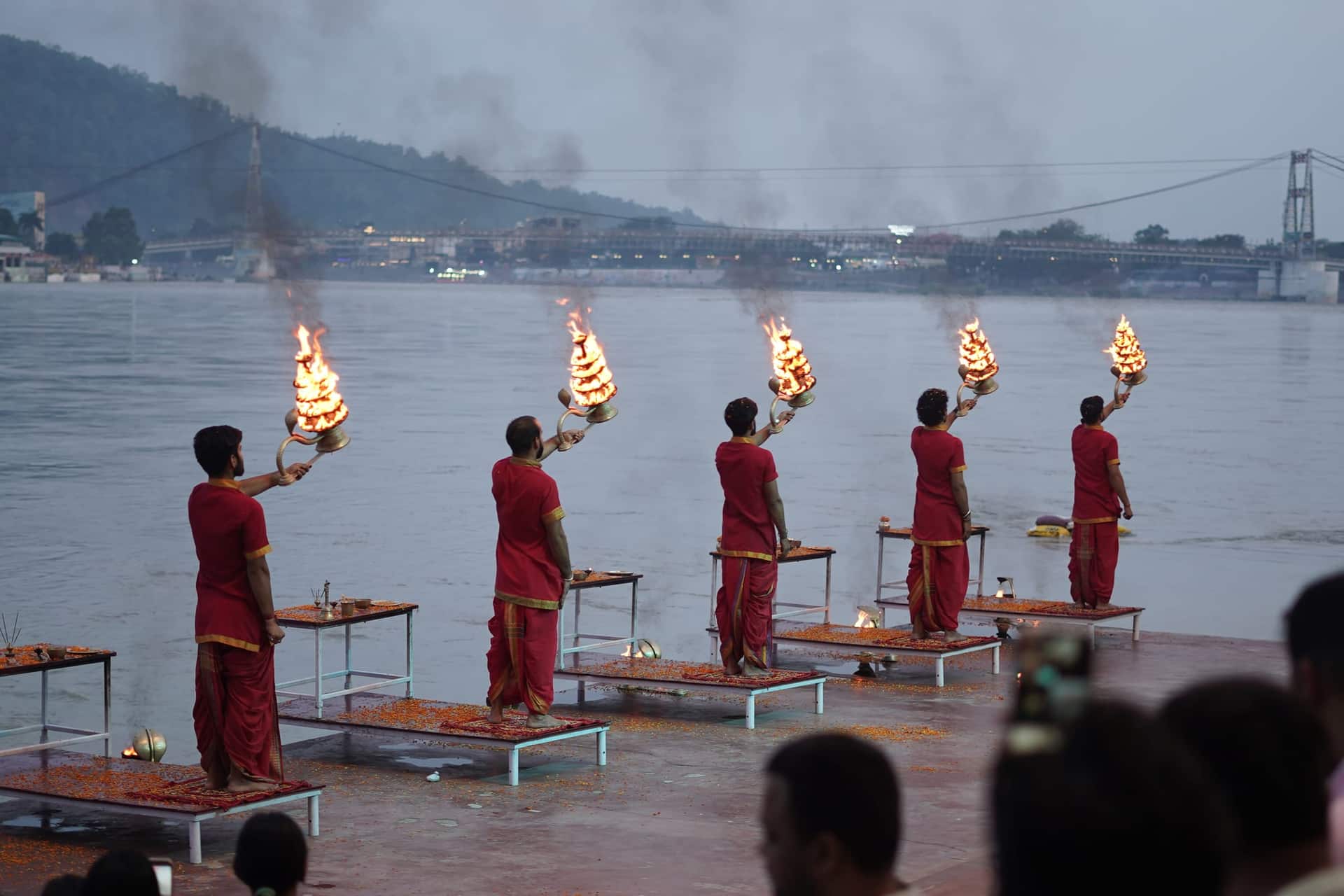 Five priests in red robes holding tiered flaming lamps during Ganga aarti on a riverside ghat in Rishikesh, with a suspension bridge visible behind.