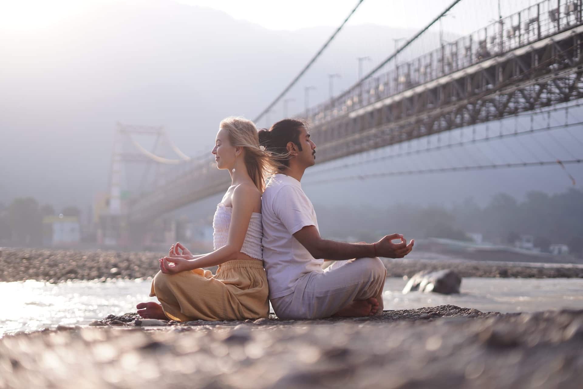 A woman and a man meditating back to back beside the Ganga river in Rishikesh, with suspension bridges behind them.
