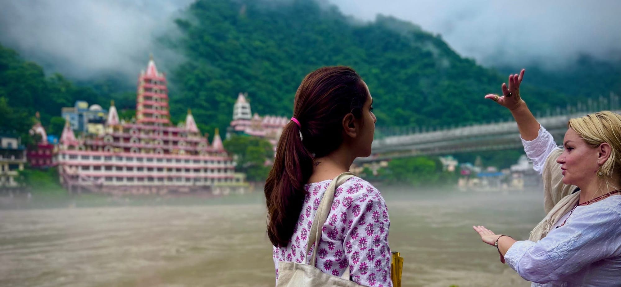 Two guests looking out over the Ganga at Trayambakeshwar Temple and Laxman Jhula bridge, one gesturing toward the view