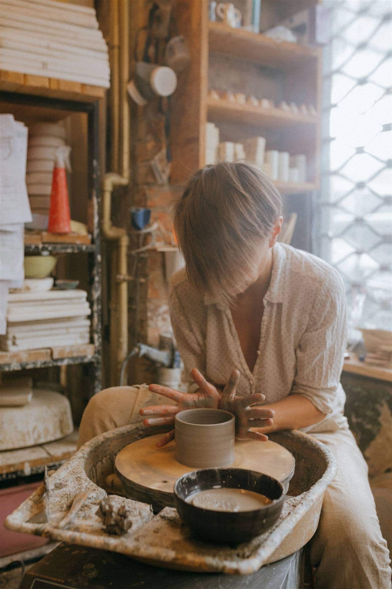 A potter leaning over a wheel, both hands shaping a wet clay cylinder, with finished pieces on the shelves behind