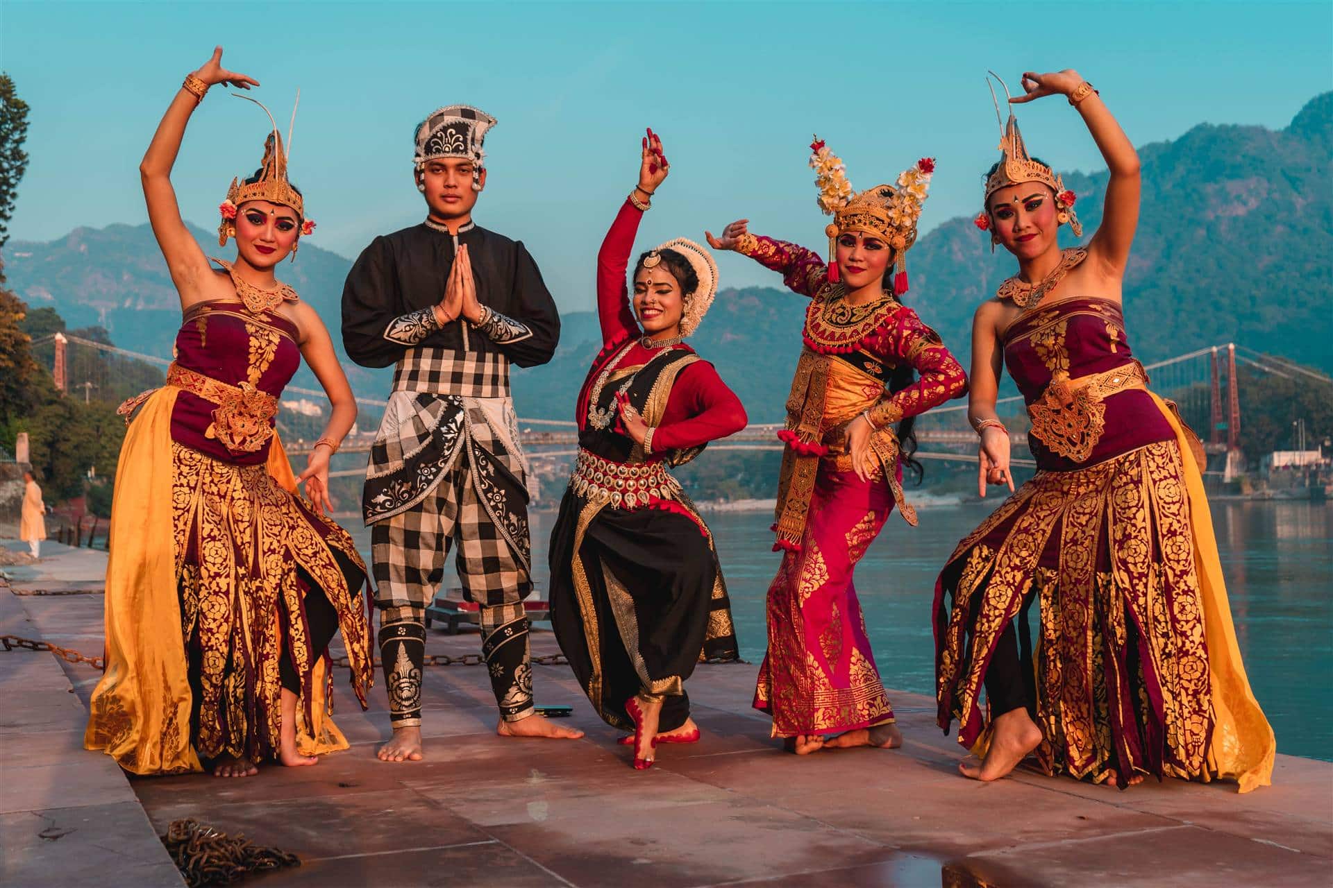 Five classical dancers in gold-embroidered costume and headdresses striking poses on a Rishikesh ghat, the Ganga and a suspension bridge behind them
