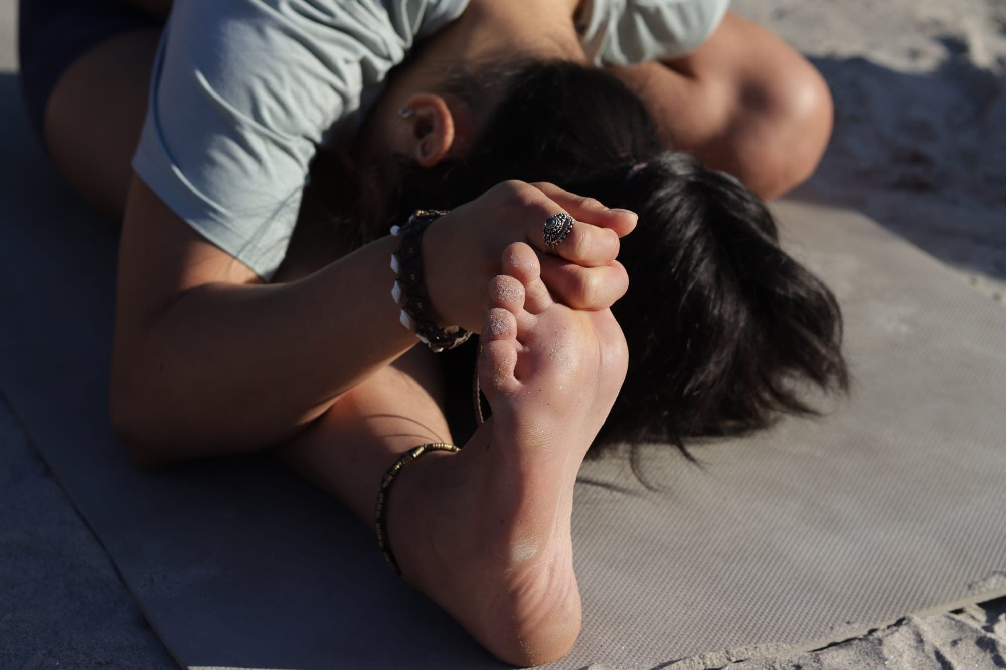 A close-up of a woman folded forward over one leg on a mat on the sand, holding the sole of her foot