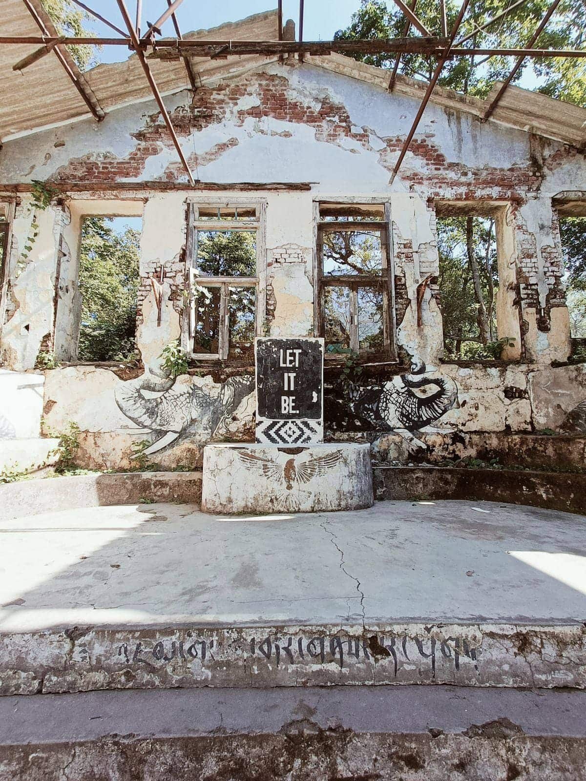A roofless brick and plaster building at the Beatles Ashram in Rishikesh, with murals and a painted LET IT BE board on the stage
