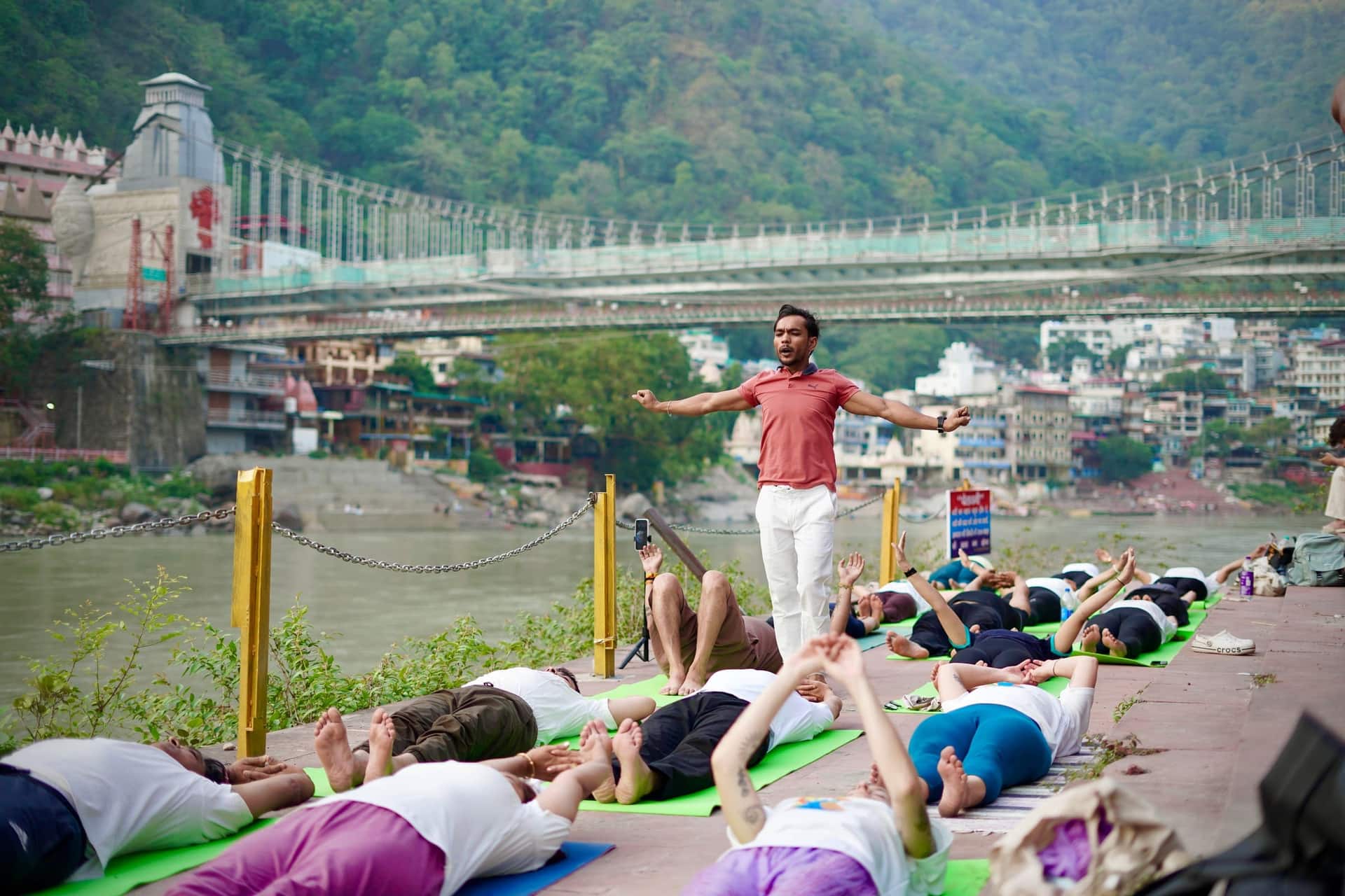 A Book My Rishikesh host leading a morning yoga class on the ghat beside the Ganga, with the Laxman Jhula bridge behind